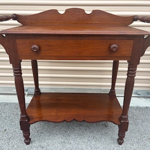 May include: A dark brown wooden side table with a drawer and a lower shelf. The table has decorative handles and legs. The table is approximately 30 inches tall, 24 inches wide, and 16 inches deep.