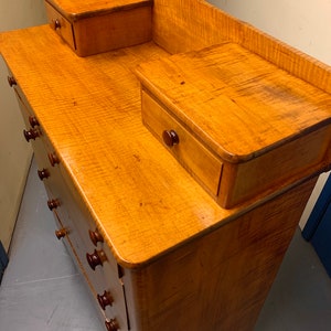 Pode incluir: Uma c&ocirc;moda de madeira clara com um padr&atilde;o de gr&atilde;o ondulado. Possui v&aacute;rias gavetas com puxadores marrons escuros e duas gavetas laterais menores. A c&ocirc;moda tem um design cl&aacute;ssico, adequada para armazenamento no quarto.