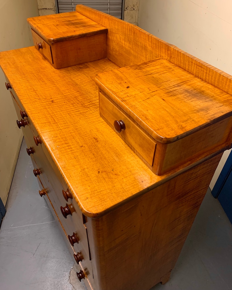 Pode incluir: Uma c&ocirc;moda de madeira clara com um padr&atilde;o de gr&atilde;o ondulado. Possui v&aacute;rias gavetas com puxadores marrons escuros e duas gavetas laterais menores. A c&ocirc;moda tem um design cl&aacute;ssico, adequada para armazenamento no quarto.
