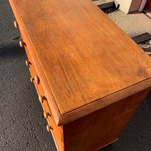 May include: A wooden chest of drawers with a light brown finish and six drawers. The drawers have wooden knobs. The chest is sitting on a black surface.