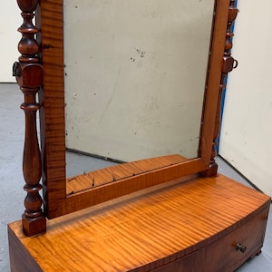 Puede incluir: Un espejo de tocador antiguo de madera con un espejo de vidrio biselado y dos cajones. El espejo está enmarcado en madera con una parte superior e inferior decorativas. Los cajones son de madera con un frente curvo y tienen pomos de madera.