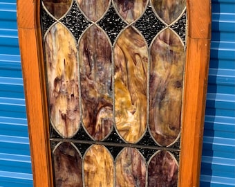 May include: An antique stained glass window with a wooden frame. The glass features a swirling pattern of amber, brown, and purple hues, divided by black lead lines. The window has an arched top and a rectangular shape.