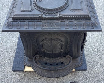 May include: A black cast iron stove with ornate detailing, including a decorative top, doors, and legs. The stove has the word "EVA" on the front. The stove is sitting on a gray mat.