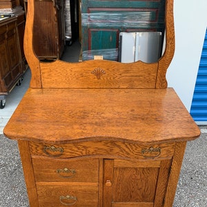 May include: Antique oak vanity with a beveled mirror, three drawers, and a single door. The vanity has ornate brass hardware and a curved top.
