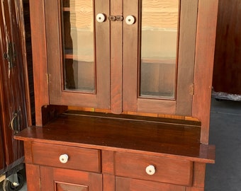 May include: A dark brown wooden cabinet with glass doors and two drawers. The cabinet has white porcelain knobs and a simple design.