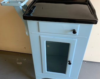 May include: A light blue metal cabinet with a black countertop, a small drawer, and a glass door. The cabinet has a metal shelf attached to the side.
