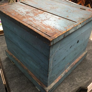 K&ouml;nnte beinhalten: Eine antike Holzkiste mit einem Scharnierdeckel. Die Kiste ist in einem verblassten Blau gestrichen und hat ein abgenutztes, rustikales Aussehen. Die Kiste besteht aus Holzbrettern und hat eine einfache, rechteckige Form.