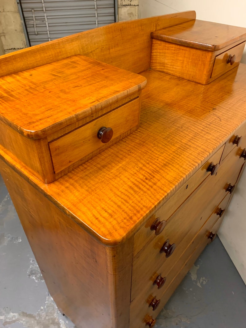 Pode incluir: C&ocirc;moda de madeira vintage com acabamento castanho claro. A c&ocirc;moda possui v&aacute;rias gavetas com puxadores castanhos escuros e dois pequenos arm&aacute;rios laterais. O veio da madeira &eacute; vis&iacute;vel e a pe&ccedil;a tem um acabamento brilhante.