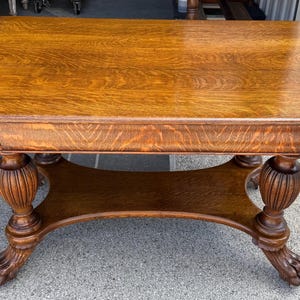 May include: A rectangular wooden table with a warm, medium-brown finish. The table features ornate, carved legs with a claw-foot design. A lower shelf connects the legs, adding to the table's functionality and aesthetic appeal. The table's dimensions are approximately 60 inches long and 30 inches wide.