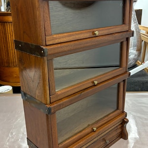 May include: A dark wood bookcase with three glass shelves and a drawer. The bookcase has brass accents and a curved top.