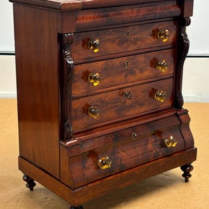 May include: A mahogany chest of drawers with five drawers and a bottom drawer with a curved front. The chest has turned legs and brass hardware. The drawers have a wood grain pattern.