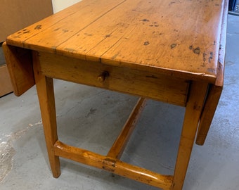 May include: A wooden drop leaf table with a single drawer. The table has a light brown finish and is made of solid wood. The table is in good condition and has a rustic look.