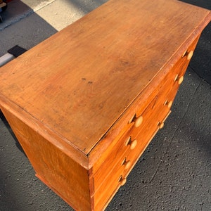 May include: A wooden chest of drawers with five drawers and simple wooden knobs. The chest is a light brown color and has a worn, rustic look.