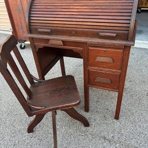 May include: An antique wooden roll-top desk with a matching chair. The desk features a curved, slatted top, two drawers, and a small cabinet. The chair has a spindle back and a swivel base. The wood has a warm, reddish-brown tone.