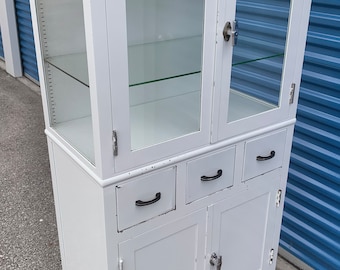 May include: White metal cabinet with glass doors and shelves. The cabinet has three drawers and two doors with metal handles. The cabinet is on wheels.