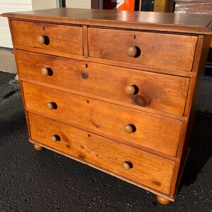 May include: A wooden chest of drawers with five drawers and rounded knobs. The chest of drawers is a light brown color and has a distressed finish.