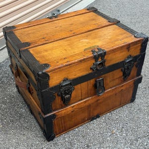 May include: A vintage wooden trunk with a light brown finish, reinforced with black metal corner protectors and bands. The trunk features ornate metal latches and a leather handle, showcasing a classic, antique aesthetic.
