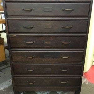 May include: A dark brown wooden dresser with six drawers. Each drawer has a brass handle and a small metal label.