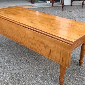 May include: A light wood drop leaf table with turned legs. The table top is made of maple wood with a natural finish. The table is in good condition and has a rustic look.