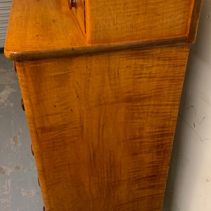 Pode incluir: Uma c&ocirc;moda de madeira vintage com acabamento castanho claro. A c&ocirc;moda tem uma frente curva com v&aacute;rias gavetas e duas gavetas pequenas no topo. O gr&atilde;o da madeira &eacute; vis&iacute;vel e o hardware &eacute; castanho escuro.