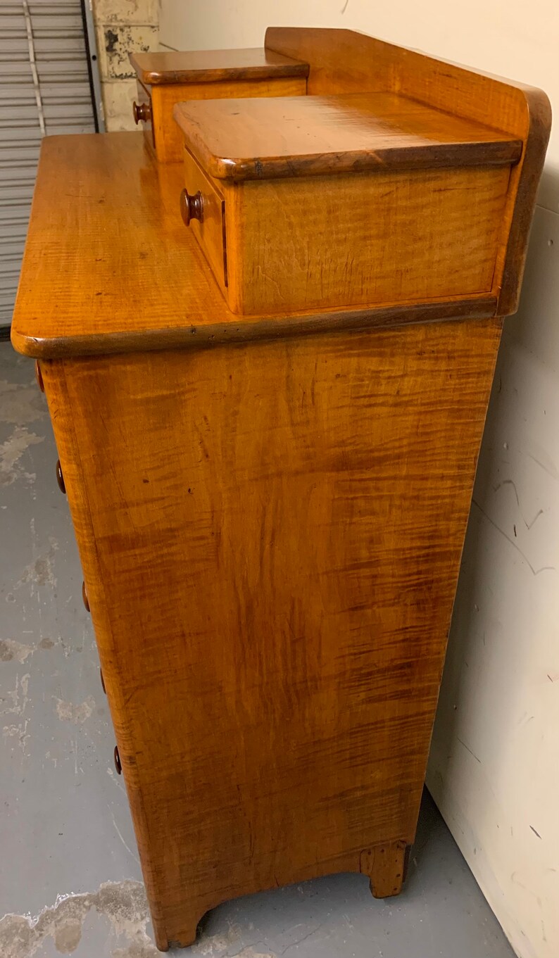 Pode incluir: Uma c&ocirc;moda de madeira vintage com acabamento castanho claro. A c&ocirc;moda tem uma frente curva com v&aacute;rias gavetas e duas gavetas pequenas no topo. O gr&atilde;o da madeira &eacute; vis&iacute;vel e o hardware &eacute; castanho escuro.