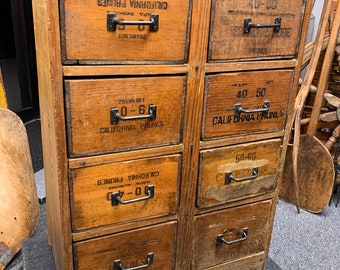 May include: A wooden cabinet with 10 drawers, each with a black metal handle. The drawers are labeled with the text "California Prunes" and "Peaches".