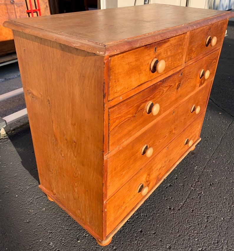 May include: A wooden chest of drawers with five drawers and wooden knobs. The chest of drawers is a light brown color and has a distressed finish.