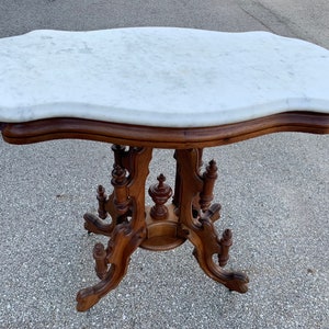 May include: A white marble top table with a carved wooden base. The table has a scalloped edge and intricate details on the base.