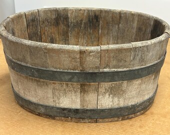 May include: A weathered, oval-shaped wooden bucket with metal bands. The bucket is made of vertical wooden planks and has a rustic, aged appearance. The metal bands are dark gray and encircle the bucket.