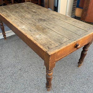 May include: A rustic wooden table with a single drawer and turned legs. The table top is made of weathered wood planks and the legs are a darker brown wood.