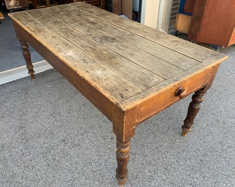 May include: A rustic wooden table with a single drawer and turned legs. The table top is made of weathered wood planks and the legs are a darker brown wood.