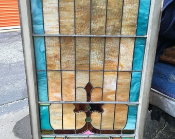 May include: A stained glass window with a geometric design featuring brown, blue, green, and pink glass. The window is framed in wood and has a weathered look.