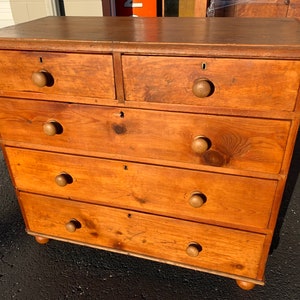 May include: A wooden chest of drawers with five drawers and a light brown finish. The drawers have wooden knobs and are slightly distressed.