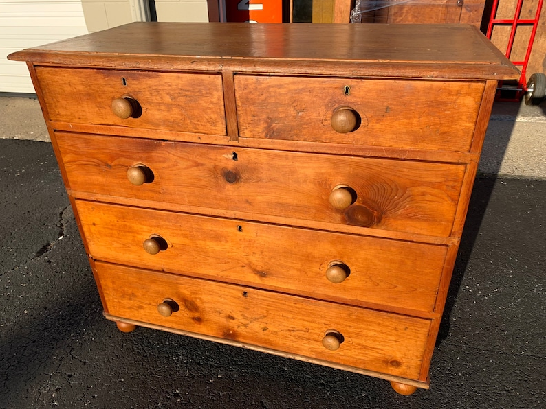 May include: A wooden chest of drawers with five drawers and a light brown finish. The drawers have wooden knobs and are slightly distressed.