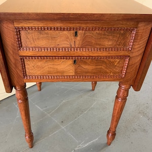 May include: A wooden drop-leaf table with two drawers and turned legs. The table is made of a rich brown wood and has a simple, elegant design.