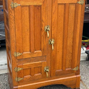 May include: An antique wooden icebox with a warm, reddish-brown finish. It features two doors with brass handles and hinges, and a small metal plaque at the top. The icebox is mounted on small wheels.