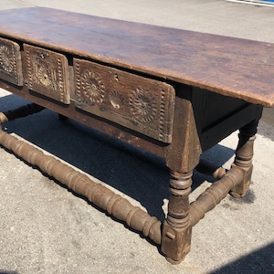 May include: A long, dark brown wooden table with three drawers. The drawers have carved floral designs. The table has turned legs and a stretcher.