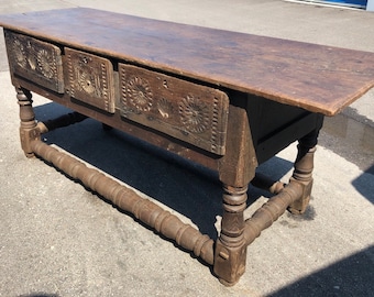 May include: A long, dark brown wooden table with three drawers. The drawers have carved floral designs. The table has turned legs and a stretcher.