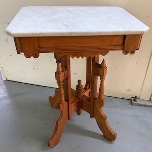 May include: A vintage marble top table with a wooden base. The table has a white marble top and a brown wooden base with intricate carvings. The base has four legs with decorative details.