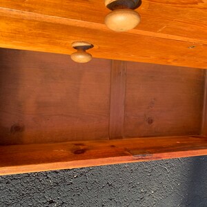 May include: A wooden chest of drawers with a single drawer open, revealing the empty interior. The chest is made of light brown wood and has simple, unadorned hardware.
