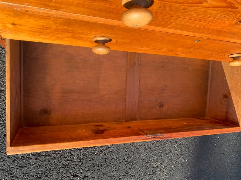 May include: A wooden chest of drawers with a single drawer open, revealing the empty interior. The chest is made of light brown wood and has simple, unadorned hardware.