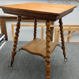 May include: An antique wooden side table with a square top and a lower shelf. The table features intricately carved spiral legs and decorative detailing around the top edge. The legs end in claw feet with small caster wheels.
