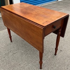 May include: A wooden drop leaf table with a single drawer and turned legs. The table is made of solid wood and has a natural finish.