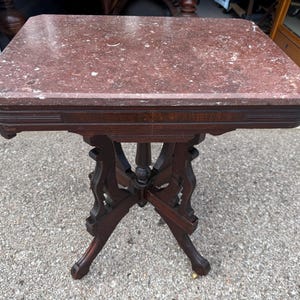 May include: A rectangular table featuring a polished reddish-brown marble top with white veining. The base is constructed from dark brown wood, showcasing a central pedestal support with four curved, outward-extending legs and a cross-brace design.