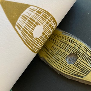 May include: A gold-colored printing block with a grid pattern and a circular hole in the center. The block is being used to create a print on a white surface.