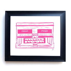May include: A pink linocut print of a storefront with the word "PRADA" printed on the windows. The storefront is framed in black.