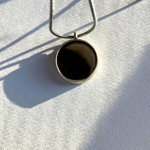 May include: A silver necklace with a circular pendant. The pendant has a dark, possibly black, center and a silver border. The chain is a thin, snake chain style. The necklace is displayed on a white surface, with shadows.