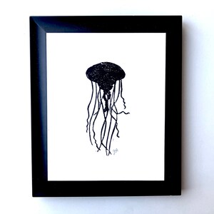 May include: Black framed art print featuring a black jellyfish illustration on a white background. The jellyfish is depicted with a textured bell and long, flowing tentacles. The artwork is signed in the lower left corner.