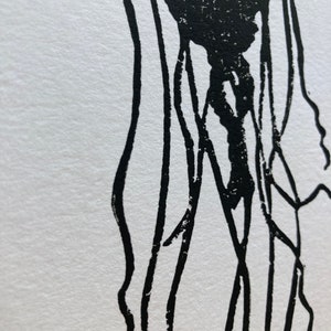 May include: A black and white linocut print of a jellyfish. The jellyfish's bell is a solid black shape, with long, thin tentacles extending downwards. The print is on white paper, creating a high-contrast image.