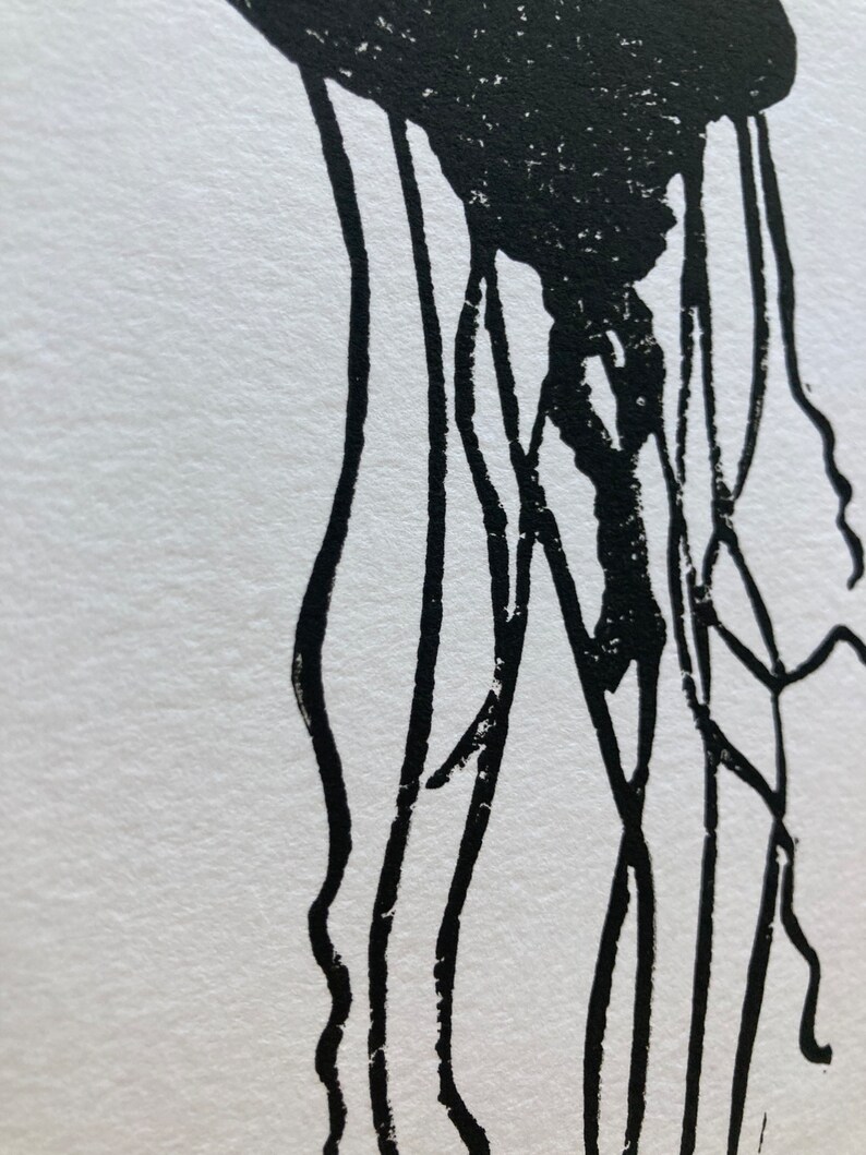 May include: A black and white linocut print of a jellyfish. The jellyfish's bell is a solid black shape, with long, thin tentacles extending downwards. The print is on white paper, creating a high-contrast image.